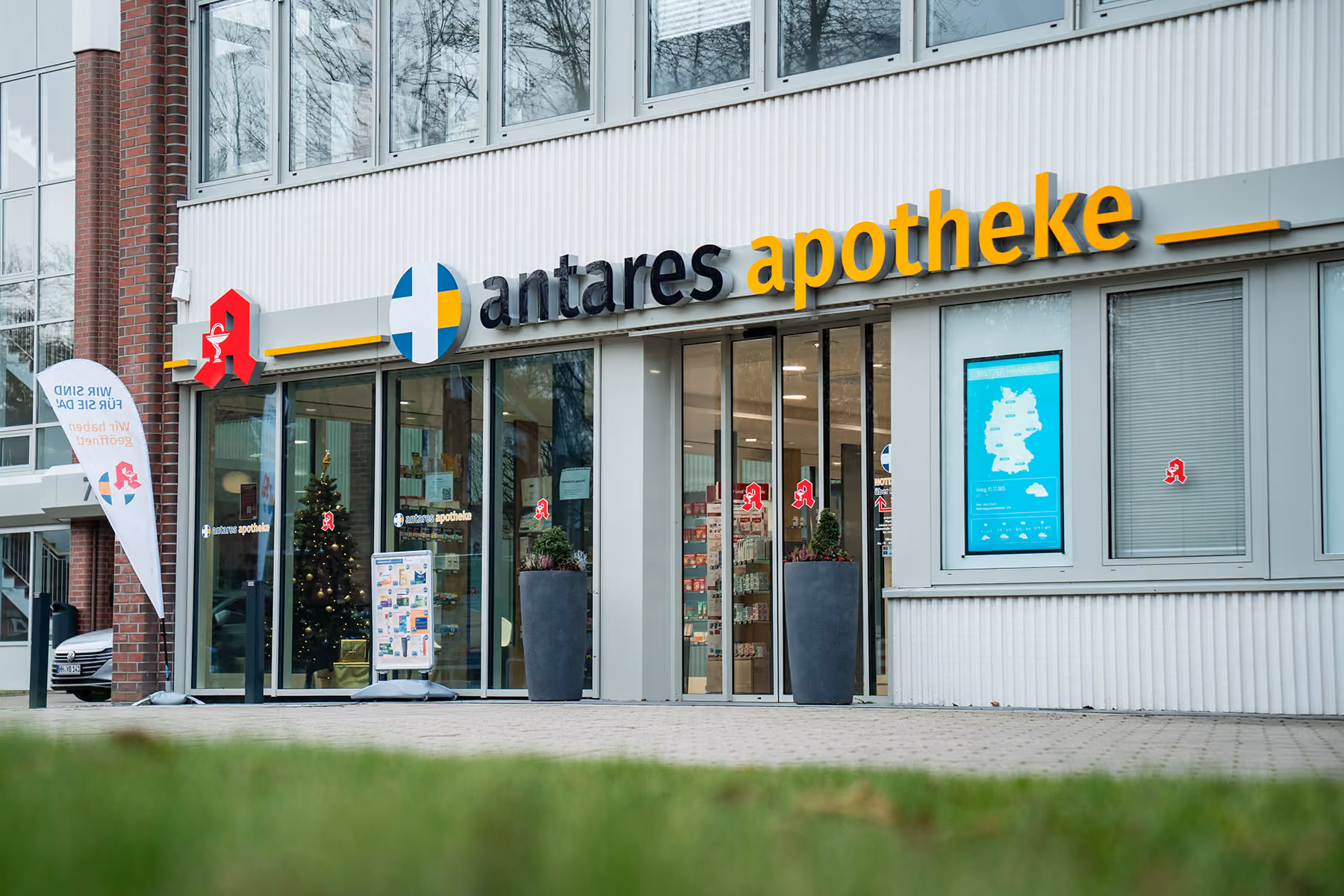 Außenansicht der Antares Apotheke mit großem Schriftzug, Glaseingang, einer Weihnachtsbaumdekoration im Inneren und Werbetafel vor dem Eingang.