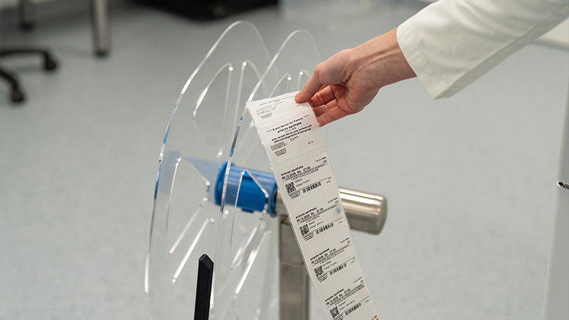 Hand in weißem Laborkittel hält einen langen, gedruckten Etikettenstreifen vor einer transparenten, runden Vorrichtung in einem Laborraum.