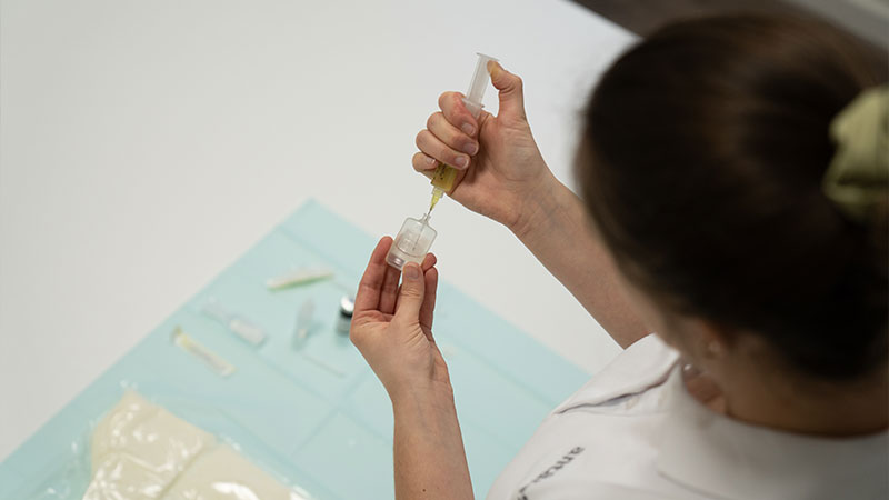 Eine Person in weißem Kittel hält eine Spritze und füllt eine Flüssigkeit in einen kleinen Behälter, auf einem Tisch liegen mehrere medizinische Utensilien und Verpackungen.