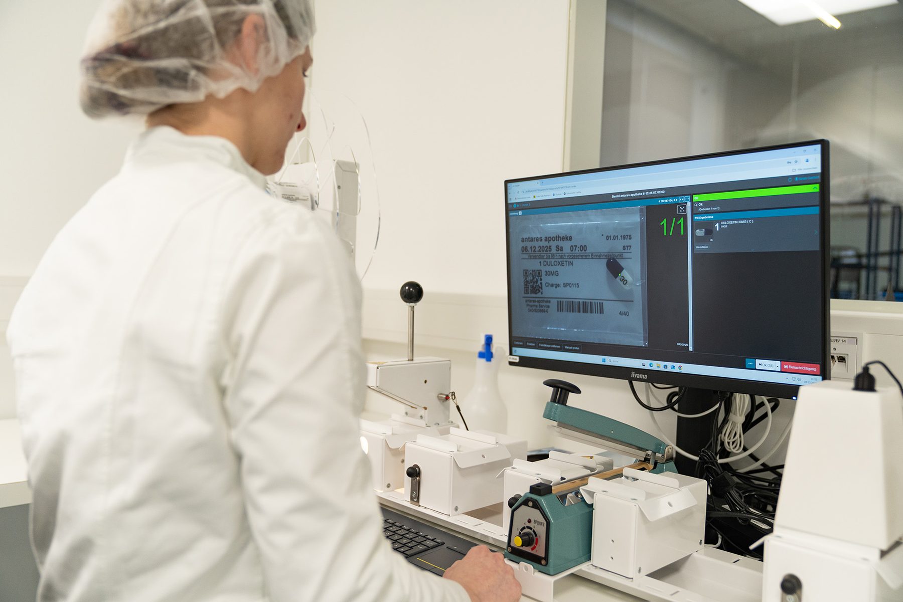 Mitarbeiterin der antares Apotheke prüft am Bildschirm die Kamerabilder eines Blisterbeutels, daneben ein Folienschweißgerät