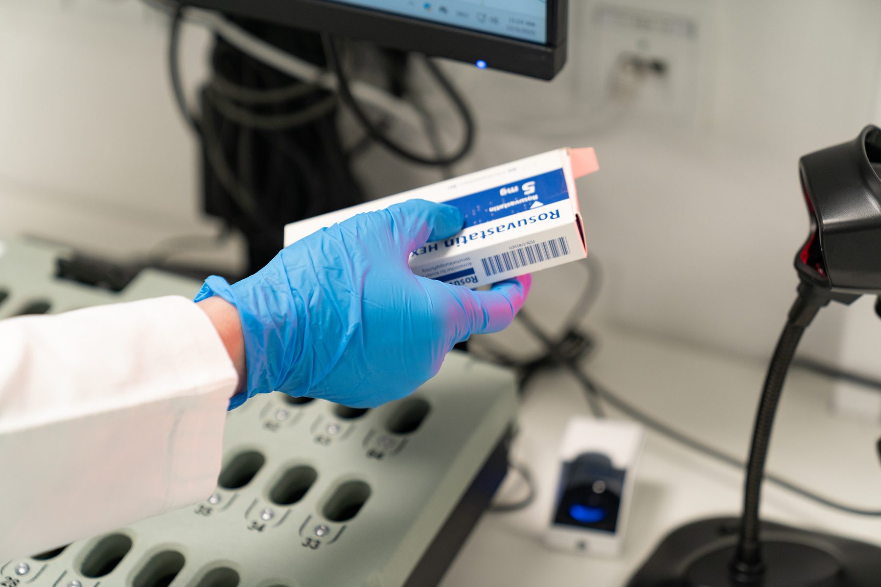 Hand im blauen Schutzhandschuh hält eine Arzneimittel-Originalpackung unter einen Barcode-Scanner, darunter ein Kassettenträger mit nummerierten Fächern